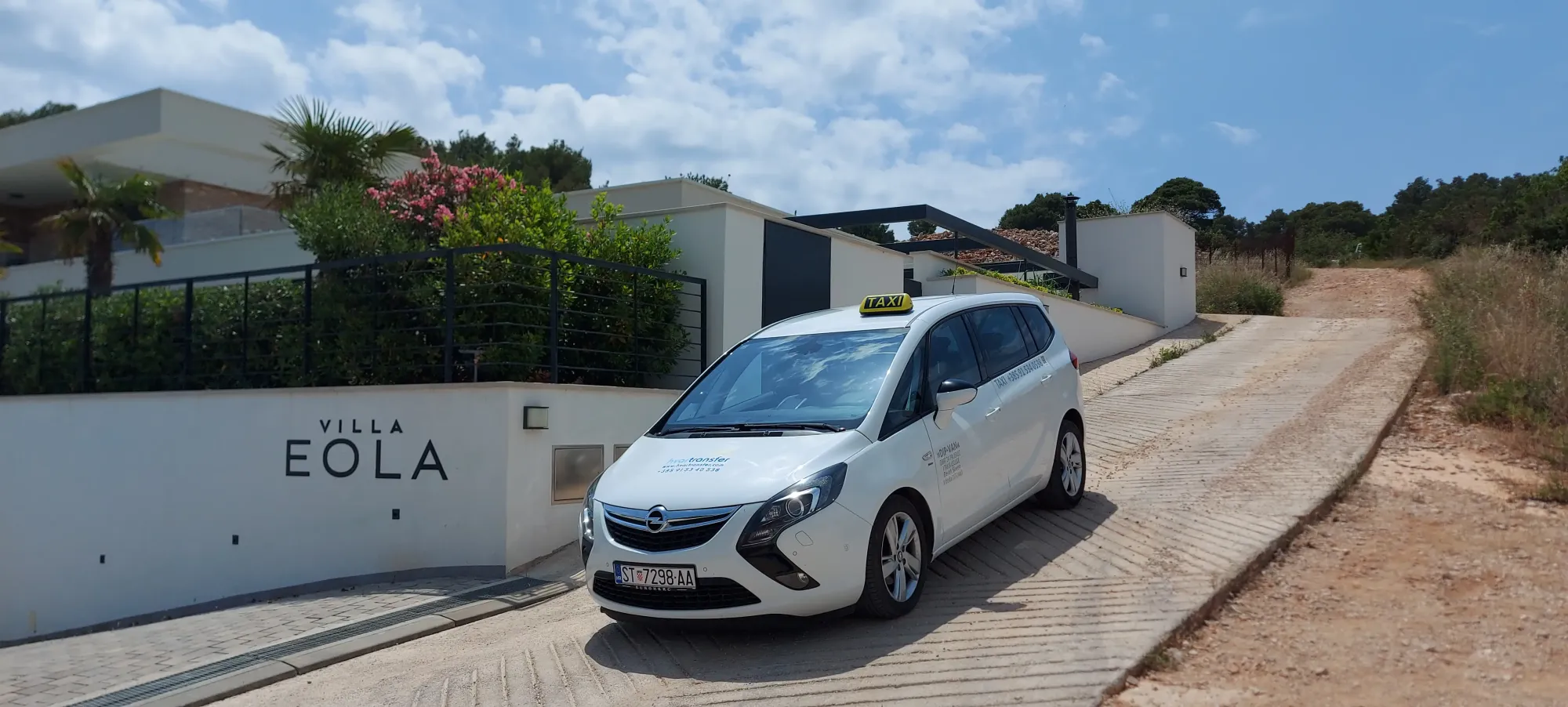 White standard taxi car parked outside Villa Eola's entrance gate on a sunny Hvar hillside