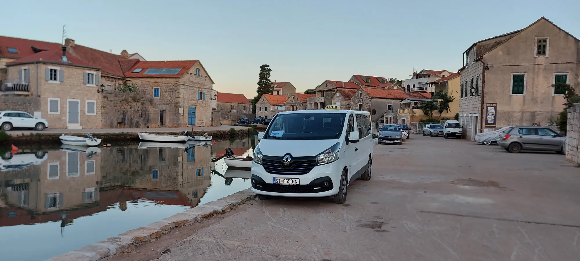 White Hvar taxi van parked on the Vrboska waterfront at dawn beside the still canal and stone houses